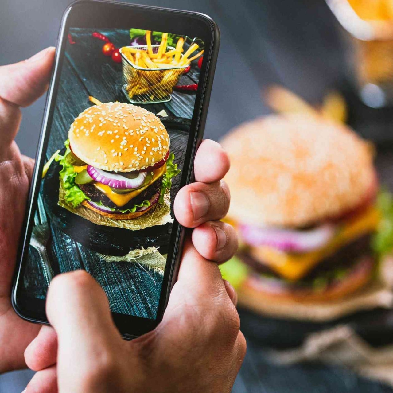 Persona saca una foto con el teléfono celular de una hamburguesa de carne con pan, lechuga, queso y cebolla, junto a una canasta de papas a la francesa. Persona saca una foto con el teléfono celular de una hamburguesa de carne con pan, lechuga, queso y cebolla, junto a una canasta de papas a la francesa.