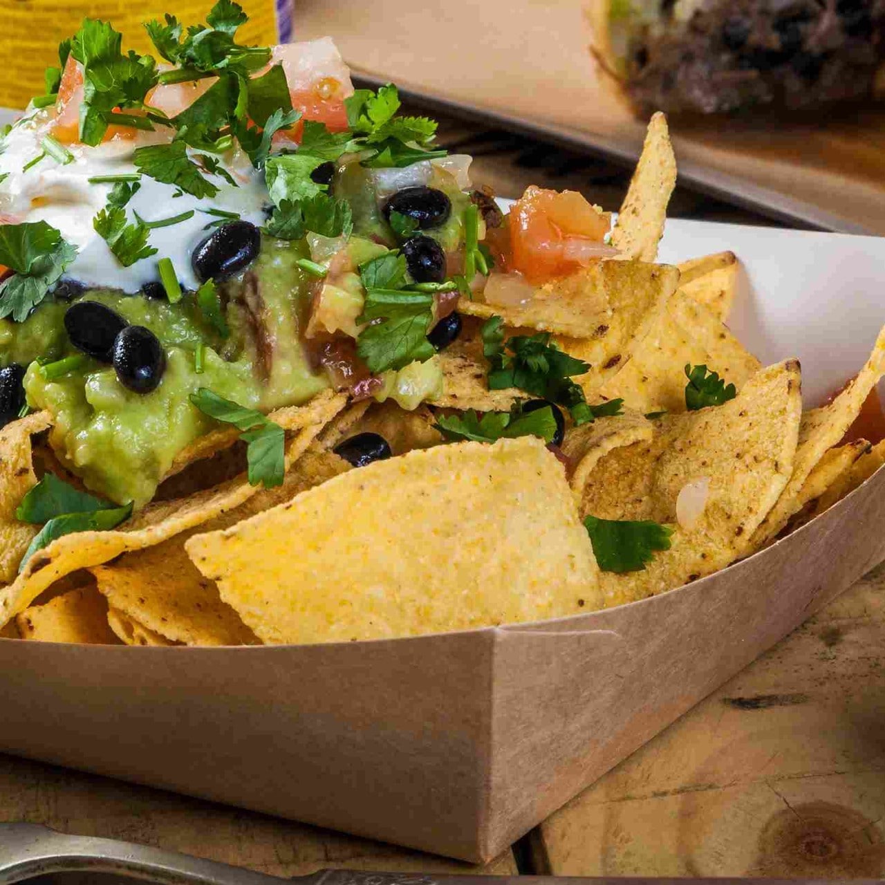 Tortillas de maíz horneadas servidas en un empaque de cartón sostenible, acompañadas de frijoles negros, guacamole, pico de gallo y cilantro.